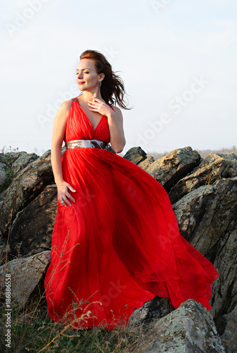 Beautiful young woman in long flying red dress standing in rocks in nature