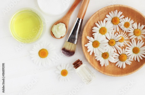 Holistic homemade herbal cosmetics with camomile, essential oils, bowls, facial cream, top view white wooden table.