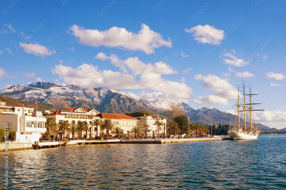 Snowy mountains and green seaside town under blue sky with white clouds.  Montenegro, winter. View of embankment of Tivat city