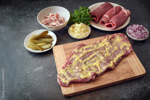 preparing beef roulades  raw meat coated with mustard and ingredients as onions  pickled cucumber and bacon on a wooden cutting board  dark kitchen countertop with copy space