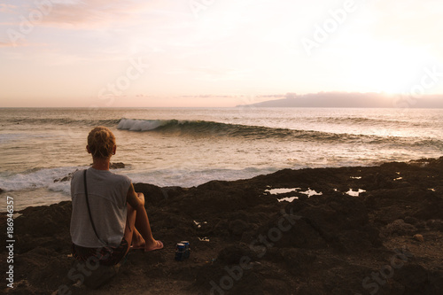 Young woman sitting on coastline at dusk with surfers on ocean waves in background while sunset