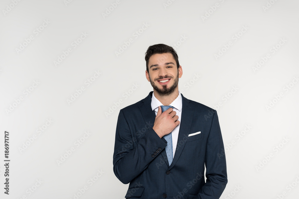 handsome young businessman adjusting necktie and smiling at camera isolated on grey handsome young businessman adjusting necktie and smiling at camera isolated on grey