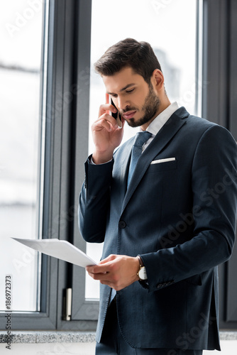 focused young businessman holding papers and talking on smartphone in office