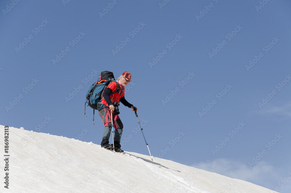a middle aged mountaineer with his bag on his back descending from a snowy hill with the help of his hiking sticks.