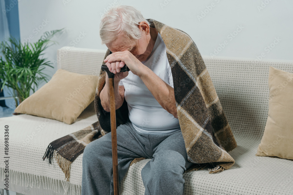 Tired senior man wearing plaid leans on a cane while sitting on sofa - obrazy, fototapety, plakaty 
