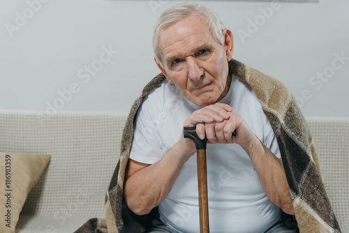 Senior man wearing plaid leans on a cane while sitting on sofa