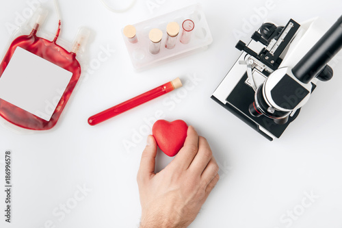 top view of hand holding red heart against vials with blood and microscope isolated on white background