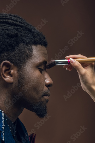 cropped image of woman applying foundation powder with makeup brush on african american man face isolated on brown