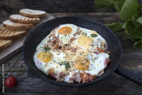 Fried eggs in an old frying pan with cheese, tomatoes, lettuce leaves and fried bread on a wooden background.