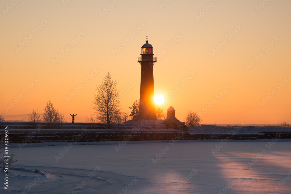 Silhouette of man on sunset background with his hands up, feeling happy and free/Silhouette of man next to a lighthouse, against the background of sunset and frozen sea/old lighthouse at Fort Kronslot