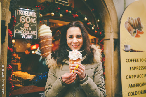 Sticker A beautiful girl in a warm jacket eats trdelnik or Trdlo with cream in her hands