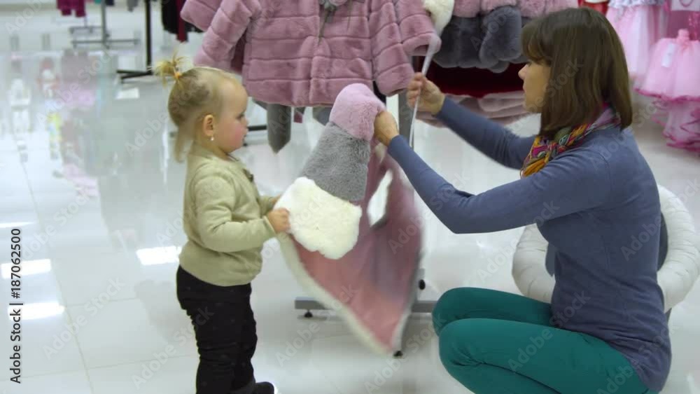 A little sweet girl with her mother chooses children's clothes in a boutique. Mom and child choose clothes in the store. A girl trying on a fur vest. A little sweet girl with her mother chooses children's clothes in a boutique. Mom and child choose clothes in the store. A girl trying on a fur vest.