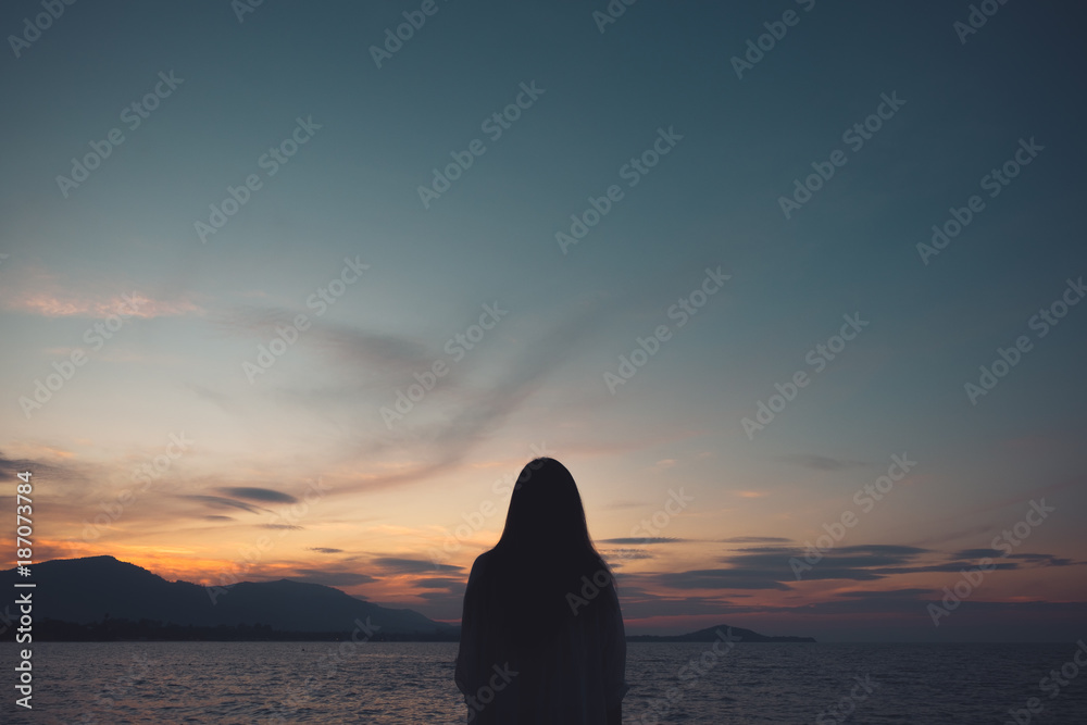 Naklejka premium Silhouettes of a woman looking at view on the beach with sea and sunset background