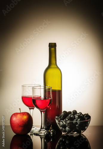 Still life with red wine / Open bottle of red wine, two glasses, an apple, and a cup with grapes standing on a table