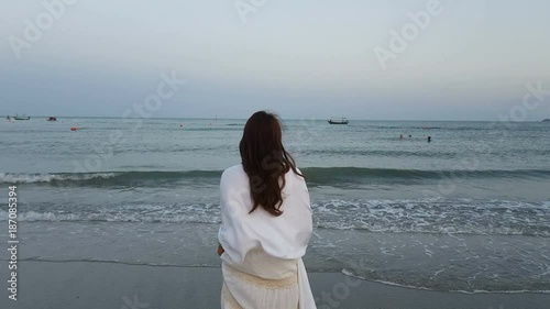 back view of young beautiful asian female standing on the beach and wind blow her hair while she is looking to the seascape and skyscape in the evening at dusk. relaxation, lifestyle, travel concept.