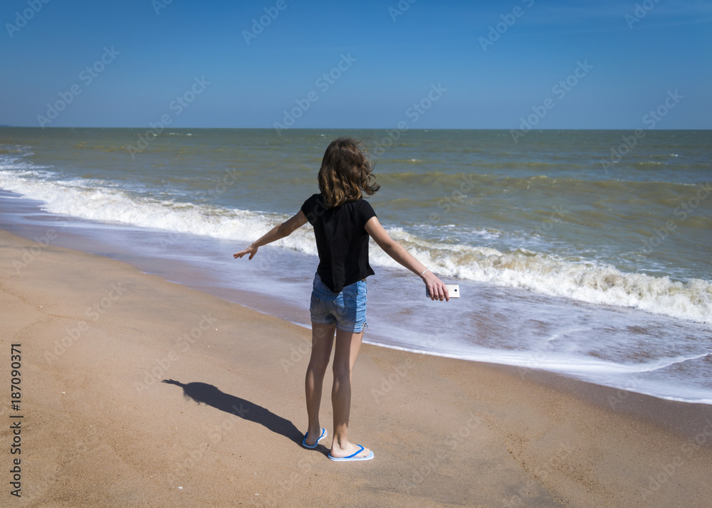 Girl stands at shore of ocean with open arms, facing water in Phuket, Thailand. Freedom, holiday concept. Teenage girl from back at seaside beach, ocean waves, enjoys the wind.