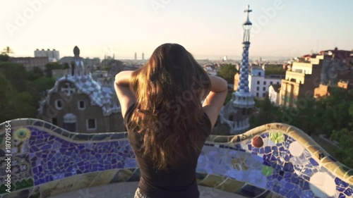 Young woman adjusting her hair while standing in Gaudi park. Beautiful girl touching hair with hand on Barcelona park at sunset. Enjoy Spain travel. Female back view near mosaic park buildings