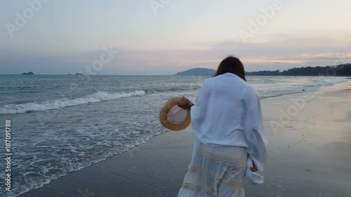 back view of young asian beautiful female in white dress walking barefoot on wet sandy beach while tide or wave is crashing to the her foots and beach when twilight or dusk.