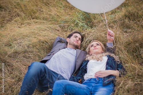 The young happy romantic couple lying in embrace on the grass outdoor and looking up and boyfriend holding the white balloons in hand