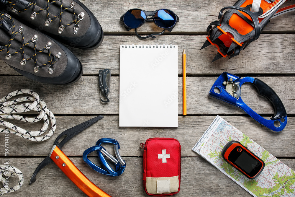 Top view of tourist equipment for a mountain trip on a rustic light wooden floor with a notebook and an empty space in the middle.