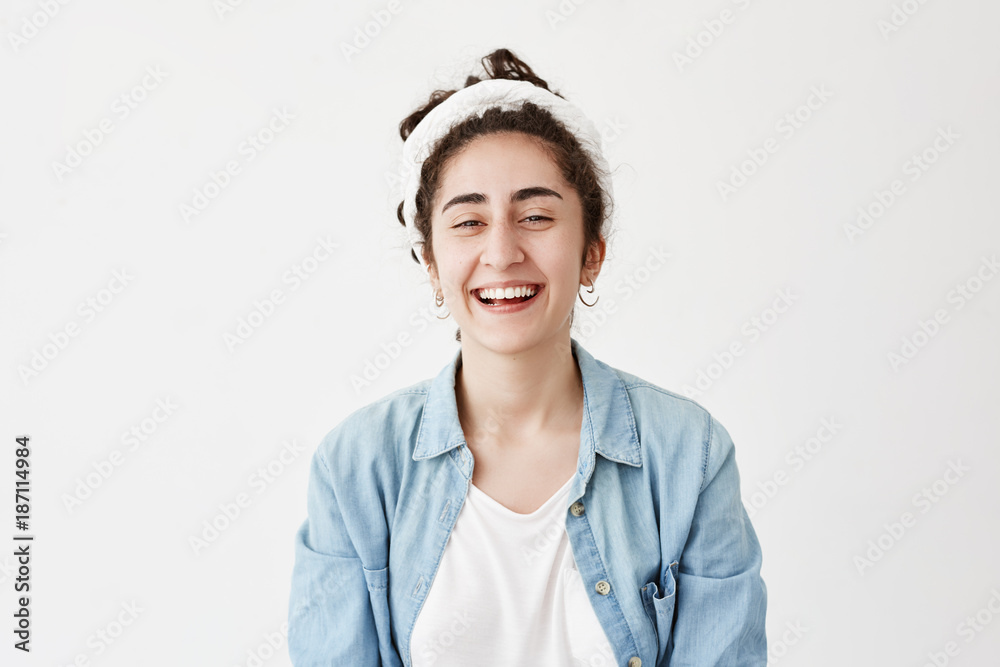 Dark-haired positive girl wears do-rag and denim shirt, looks happily at camera, smiles broadly, laughs at joke, rejoices youth. Female model poses against white backgorund demostrates white teeth