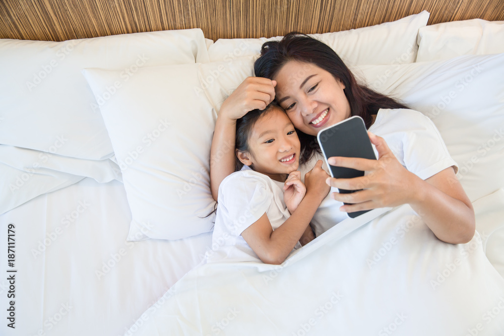 Asian mother and daughter using mobile phone on white bed in morning time Asian mother and daughter using mobile phone on white bed in morning time