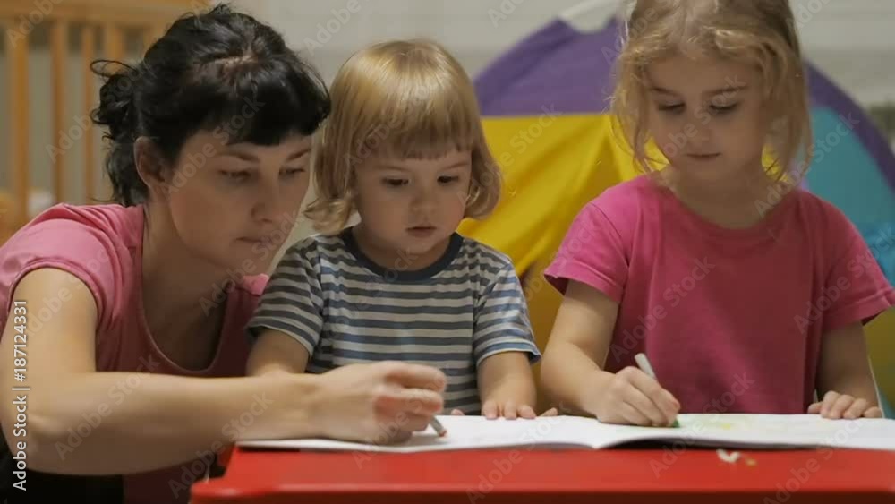 The educator and two little children girl and boy sitting at table drawing. Brother and sister and their mother drawing with colorful crayons and markers The educator and two little children girl and boy sitting at table drawing. Brother and sister and their mother drawing with colorful crayons and markers