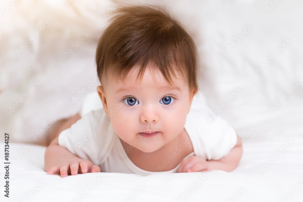 Smiling little baby girl with big blue eyes lying on her tummy on white bed looking at camera and smiling