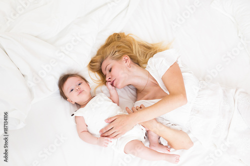 Mother and child on a white bed top view. Mom and baby girl with blue eyes playing in white bedroom. Parent and little kid relaxing at home and having fun together