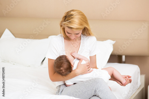 A young mother breastfeeding her little baby girl while holding in her arms at light spacious domestic bedroom
