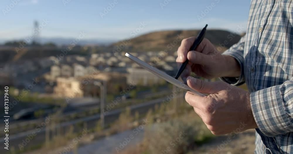 Developer or architect hands using tablet computer with stylus. Buildings under construction in distant rural suburb background. Screen not visible, 4K.