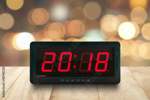 red led light illuminated numbers 2018 on digital electric alarm clock face on brown wooden table top with defocused colorful Christmas lights bokeh background, time symbol for beginning New Year