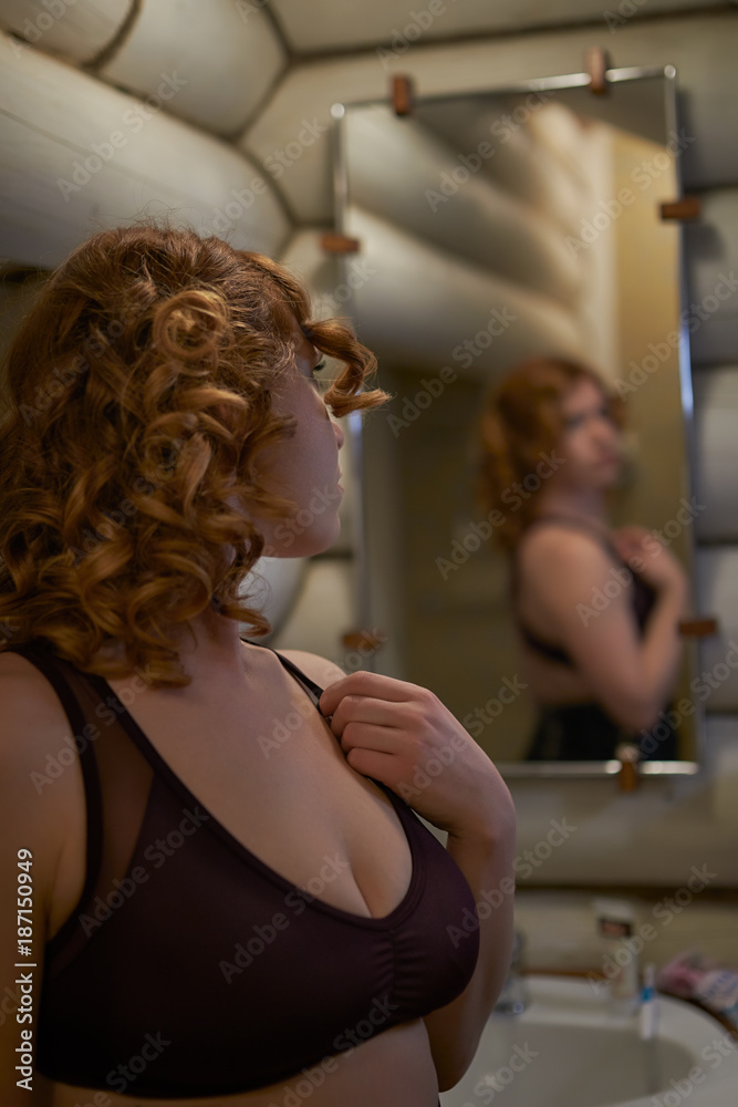 Fototapeta premium Redheaded young woman looking at herself in the mirror in the bathroom