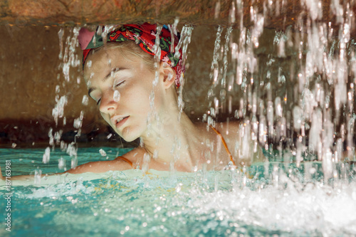 Portrait of a beautiful blonde young woman captured in swimming suit in a secret water cave. Her body is in the water  in front of her is waterfall and she enjoys the sun s rays on her soft skin.