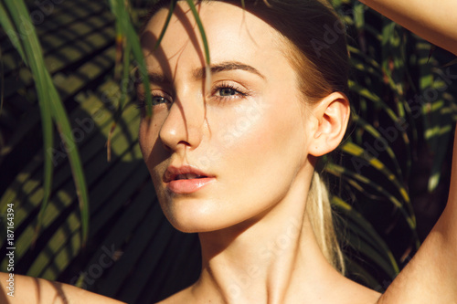 Close-up portrait of beautiful blonde young woman with perfect skin, beautiful green eyes and long neck hiding under palm leaf