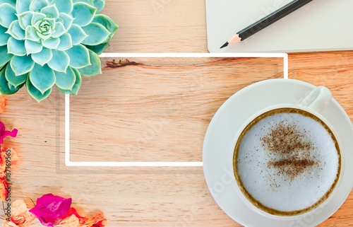 Cappuccino coffee in a white cup with blurry confection and blurry red Flower on wooden table with white frame and Cactus for design background