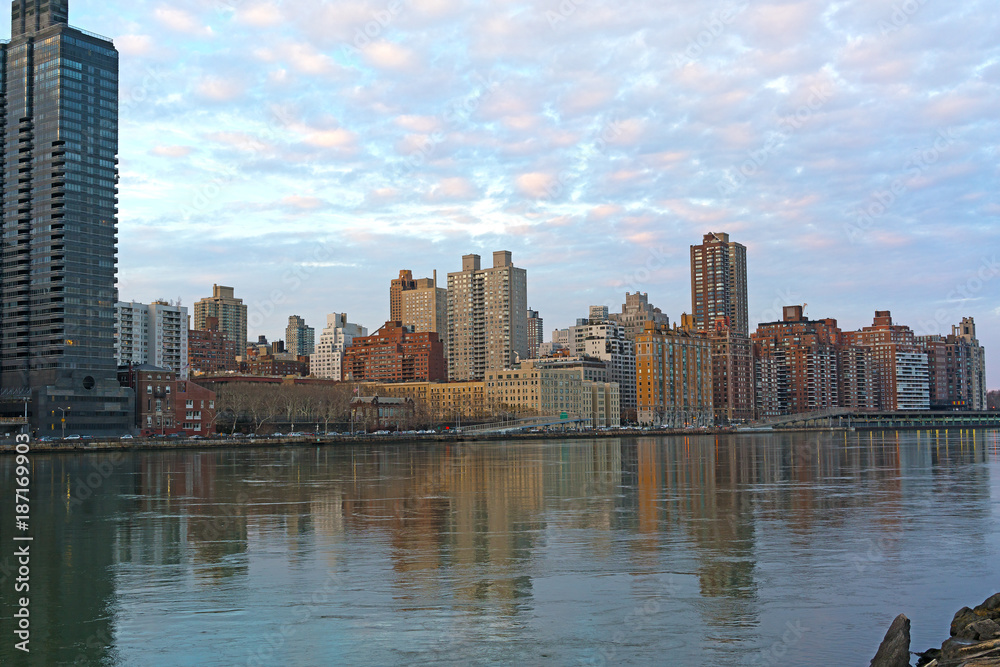 Obraz premium Urban landscape at sunset in winter along East River in Manhattan, New York. Buildings reflection in harmony with pastel hues of cumulus clouds during the quiet city sunset.