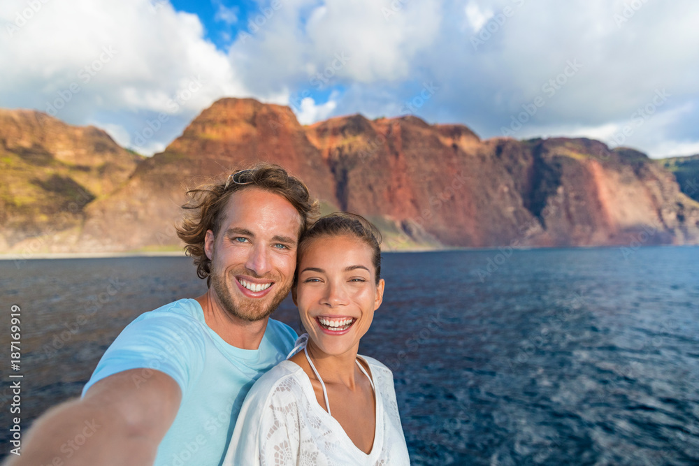 Obraz premium Happy couple tourists on sunset cruise in Na Pali Coast Kauai, Hawaii taking selfie photo with mobile phone app. Smiling Asian woman and Caucasian man taking picture on summer holiday.