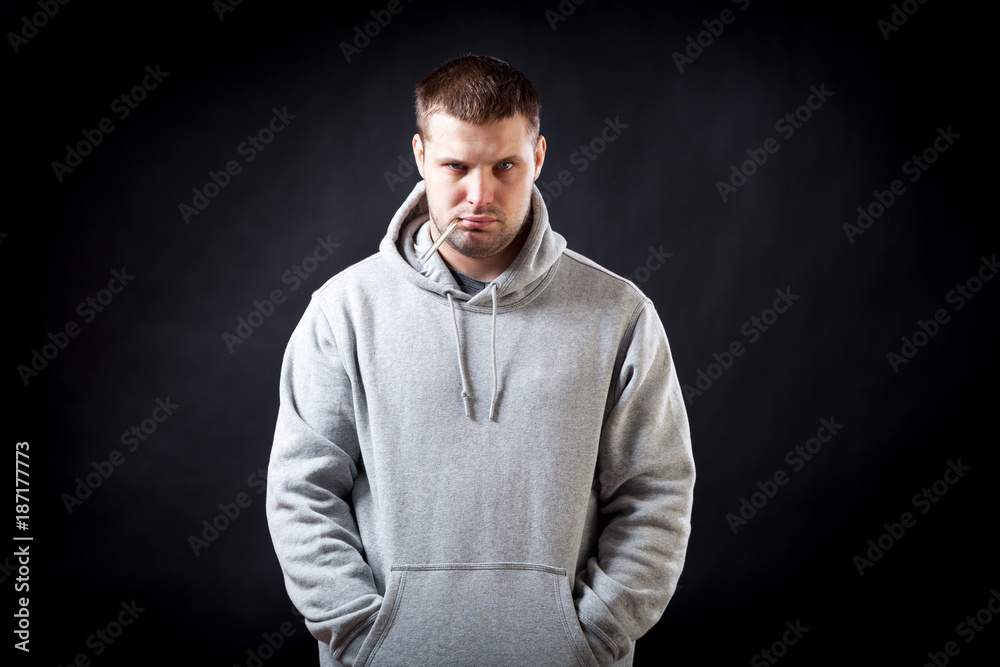 A young, dark-haired, sad-looking man in a sporty gray sweatshirt fell ill with a cold, measured the temperature on a black isolated background. A man holding a thermometer in his mouth