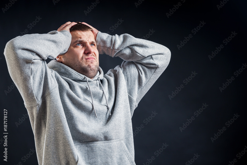 A young dark-haired man in a sporty gray sweatshirt clings to his head, which hurts very much on a black isolated background, the torturer suffers from a headache
