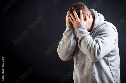 A young dark-haired man in a sporting gray sweatshirt is upset, upset and keeps his hands on his head on a black isolated background, the torturer suffers a headache