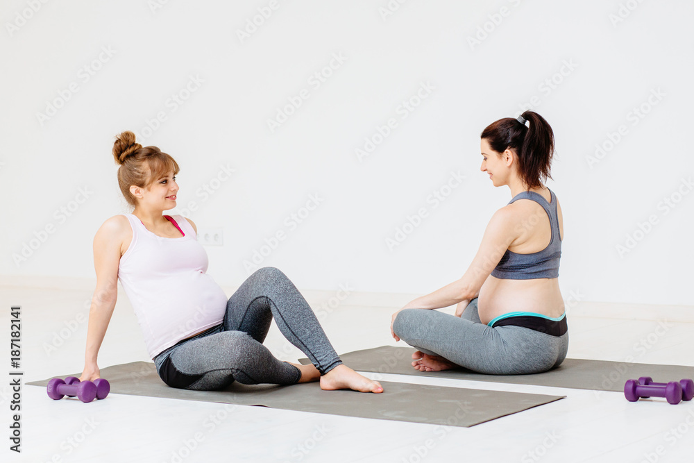 Two pregnant female in gray sportswear relaxing and talking after workout while sitting on gimnastic mats in yoga class. Two pregnant female in gray sportswear relaxing and talking after workout while sitting on gimnastic mats in yoga class.