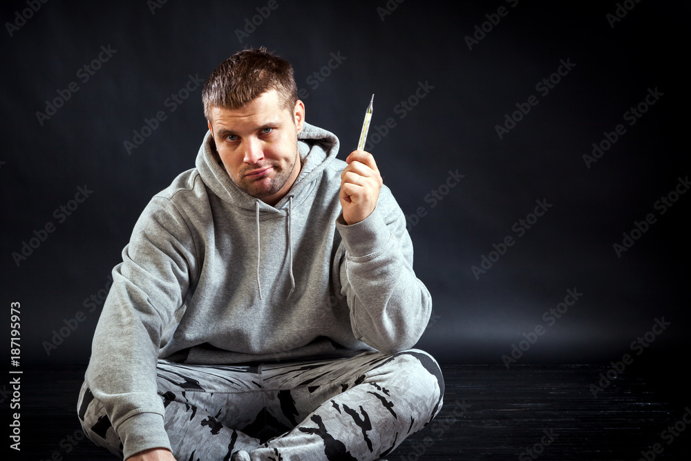 A young sad man in sports clothes fell ill with a cold, measured the temperature with a thermometer and was upset on a black isolated background. A young sad man in sports clothes fell ill with a cold, measured the temperature with a thermometer and was upset on a black isolated background.