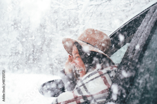 Happy woman is wearing hat and checkered scarf hanging out of the car window traveling on winter forest and enjoying heavy snowfall. Smiling hipster girl admire snowy road