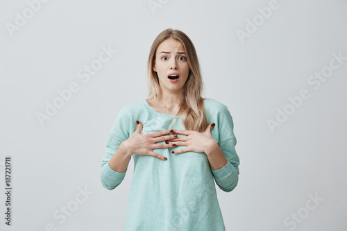 Surprised terrified blonde female gestures with uncertainty, stares at camera, puzzled as doesn`t know anwser on question, isolated against gray background. People, body language, emotions concept photo