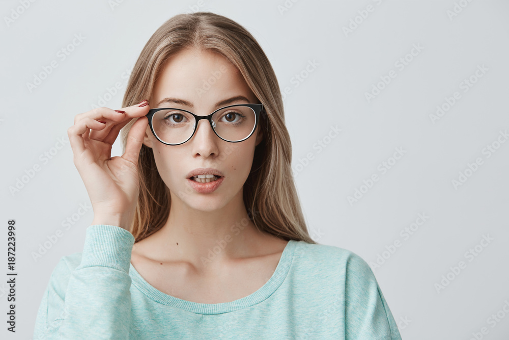 Delightful pretty blonde woman in trendy eyeglasses, wears light blue sweater, stands indoors, looks at camera with appeal. Lifestyle, beauty and youth concept.