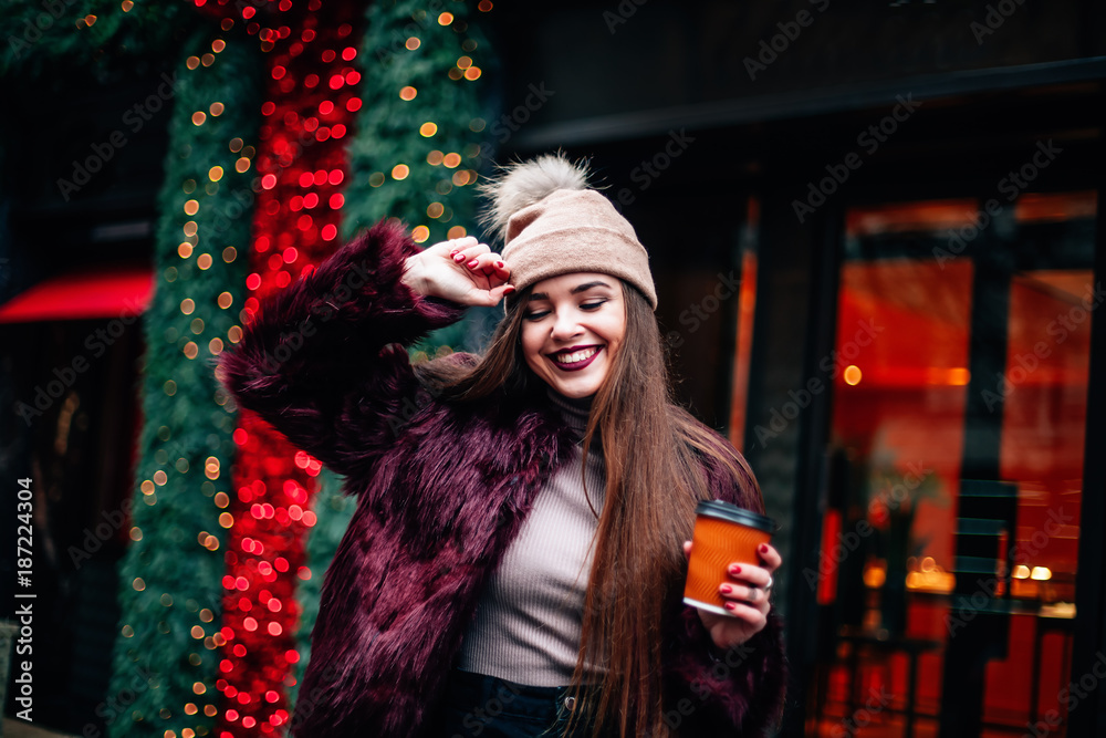 The concept of street fashion. portrait of Young girl dressed in a fashionable outfit. Posing against the window of the boutique girl smiling and drinking coffee