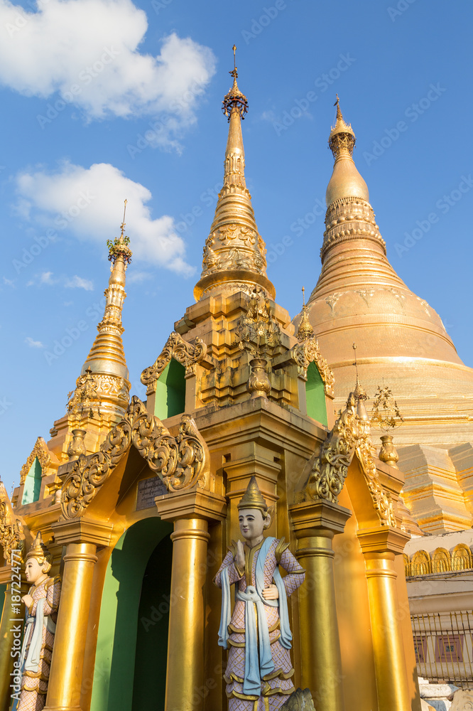 Two small pagodas and statues in front of the gilded Shwedagon Pagoda in Yangon, Myanmar on a sunny day.