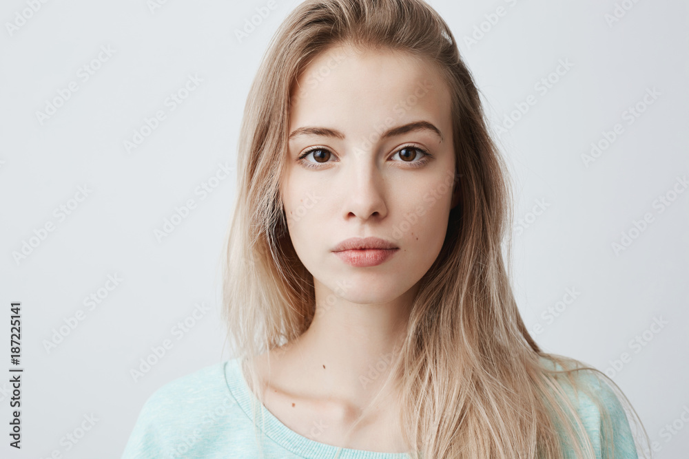 Obraz premium Jasnowłosa kobieta o ciepłych ciemnych oczach, zdrowej skórze patrząc w kamerę z zadowolonym i spokojnym wyrazem. Powabna dziewczyna o pięknym wyglądzie ubrana niedbale pozuje w studio. Pojęcie piękna