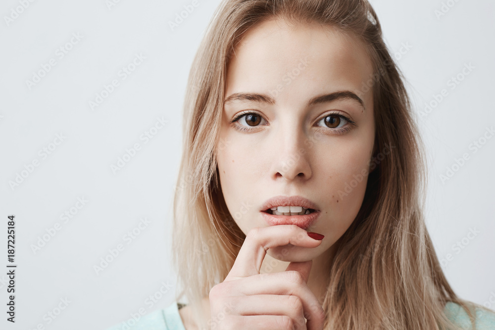European beautiful young female with blonde dyed straight hair looks at camera with interest, has pensive expression of face, makes decision, holds finger on chin, isolated against gray background. European beautiful young female with blonde dyed straight hair looks at camera with interest, has pensive expression of face, makes decision, holds finger on chin, isolated against gray background.